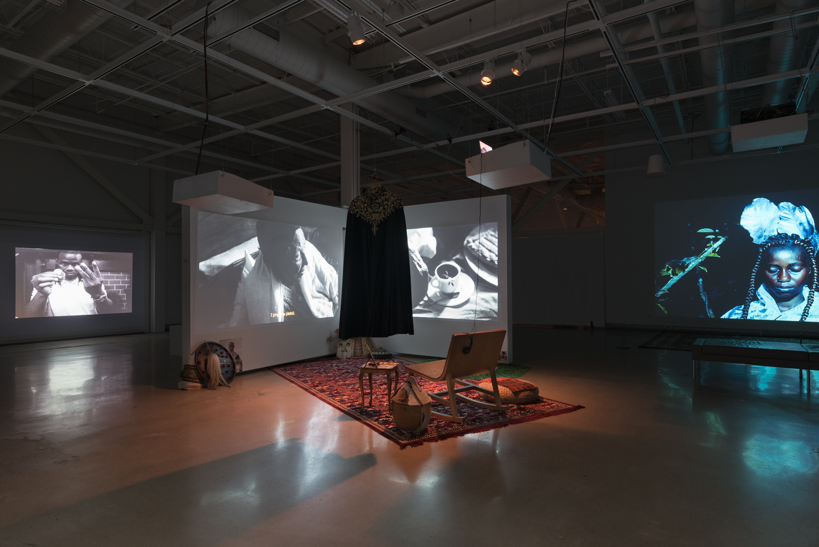 A photo of a gallery exhibition featuring multiple projected videos on white screens, plus a carpeted viewing area in the middle of an otherwise dark room.