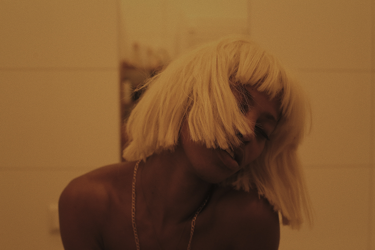 A still image from Rikki Wright's "A Song About Love" shows a Black person in a blonde wig, seemingly in a state of ecstasy or mid-dance, their head tilted to the side and hair obscuring much of their face.