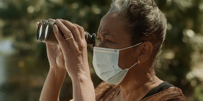 An older Black person looks through a pair of binoculars while fairing a surgical face mask, trees out of focus in the distance behind them.