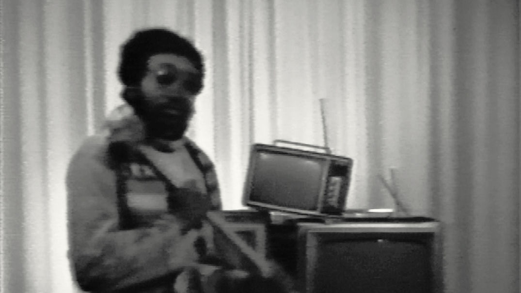 A blurry black-and-white photo of Ulysses Jenkins from his Mass of Images (1978) video. Ulysses is wearing sunglasses and is facing the camera. Behind him is a set of antenna TVs.
