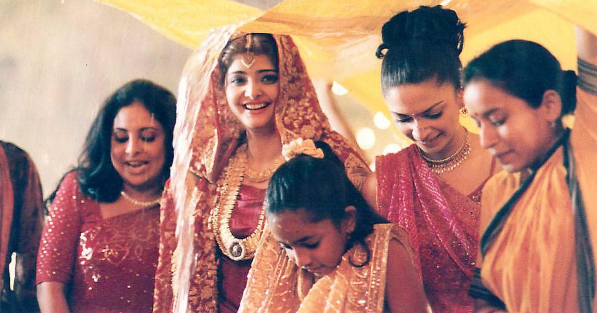 A group of wedding goers gather under a tent during a storm, they are dressed glamorously in reds and marigolds, with one person at the center appearing to be the bride.