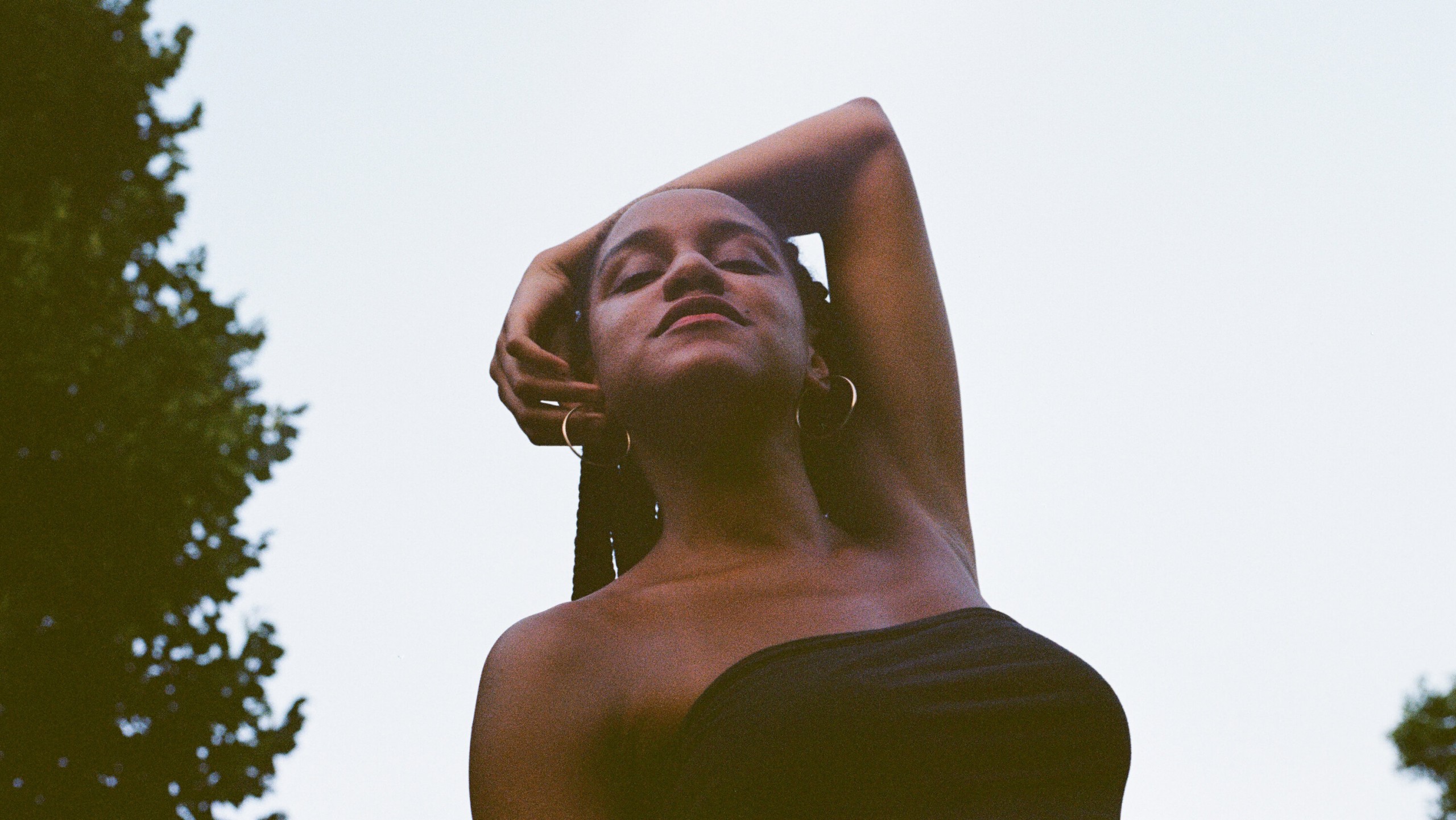 A still from Beba. A portrait of a Black Latinx woman. She has a peaceful expression, her eyes are closed and her arm is draped over her head. Behind her is the open sky.