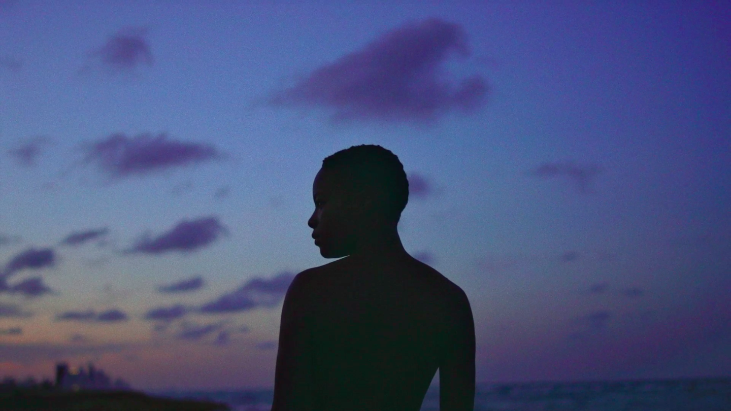 A Black person stands silhouetted against an early evening sky - the sun setting in the far distance. They look to their left.