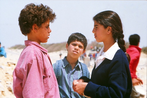 A film still from "The Tale of the Three Jewels". It shows three young people standing together outside. A young boy and young girl are facing one another. They appear to be in a serious discussion. There is a third young boy in the middle looking at them.