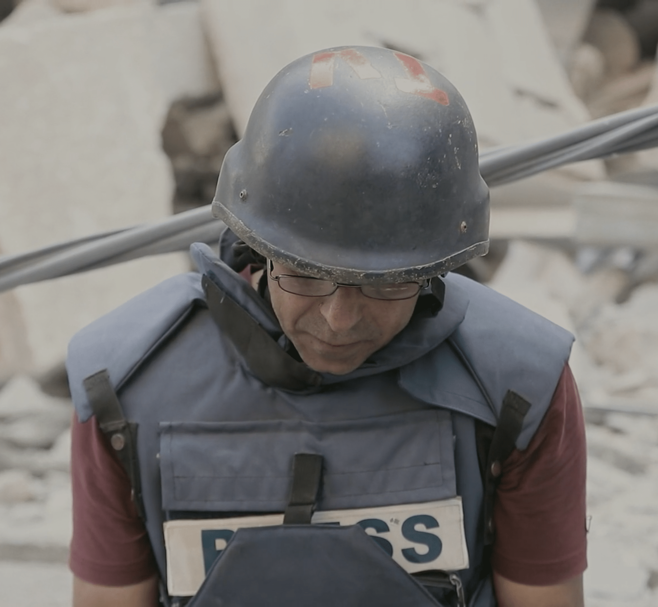 A still from Bank of Targets, it’s a close-up shot of a man wearing a press vest, designed for journalists operating in active war zones, and a helmet. He is standing in front of a pile of rubble.