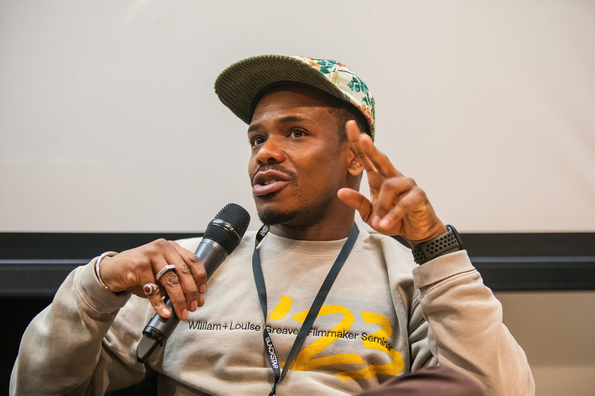 A Black person wearing a baseball hat is speaking into a microphone while wearing a grey sweatshirt.