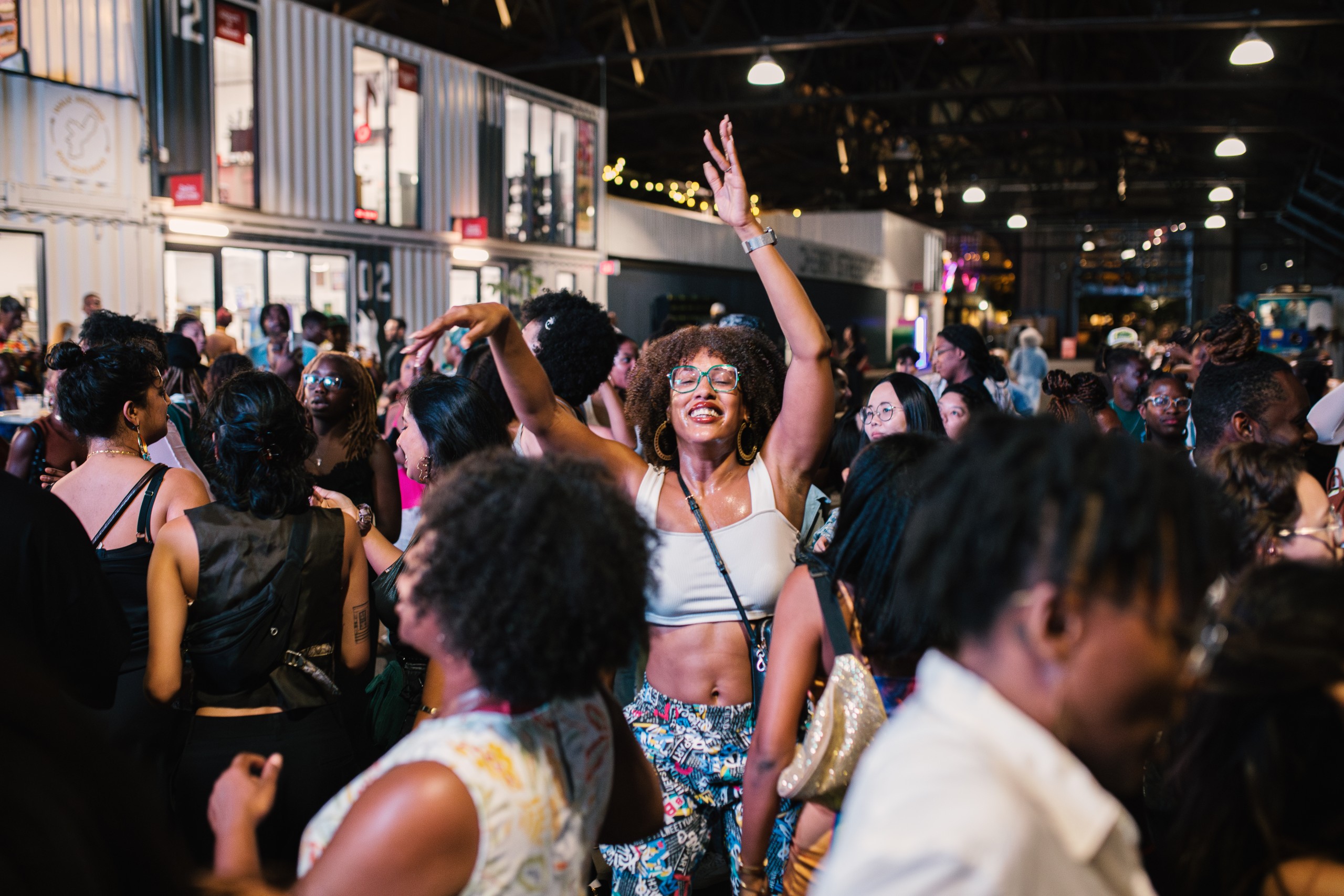 A photo of a person dancing with their arms raised, surrounded by other Black and brown people dancing.