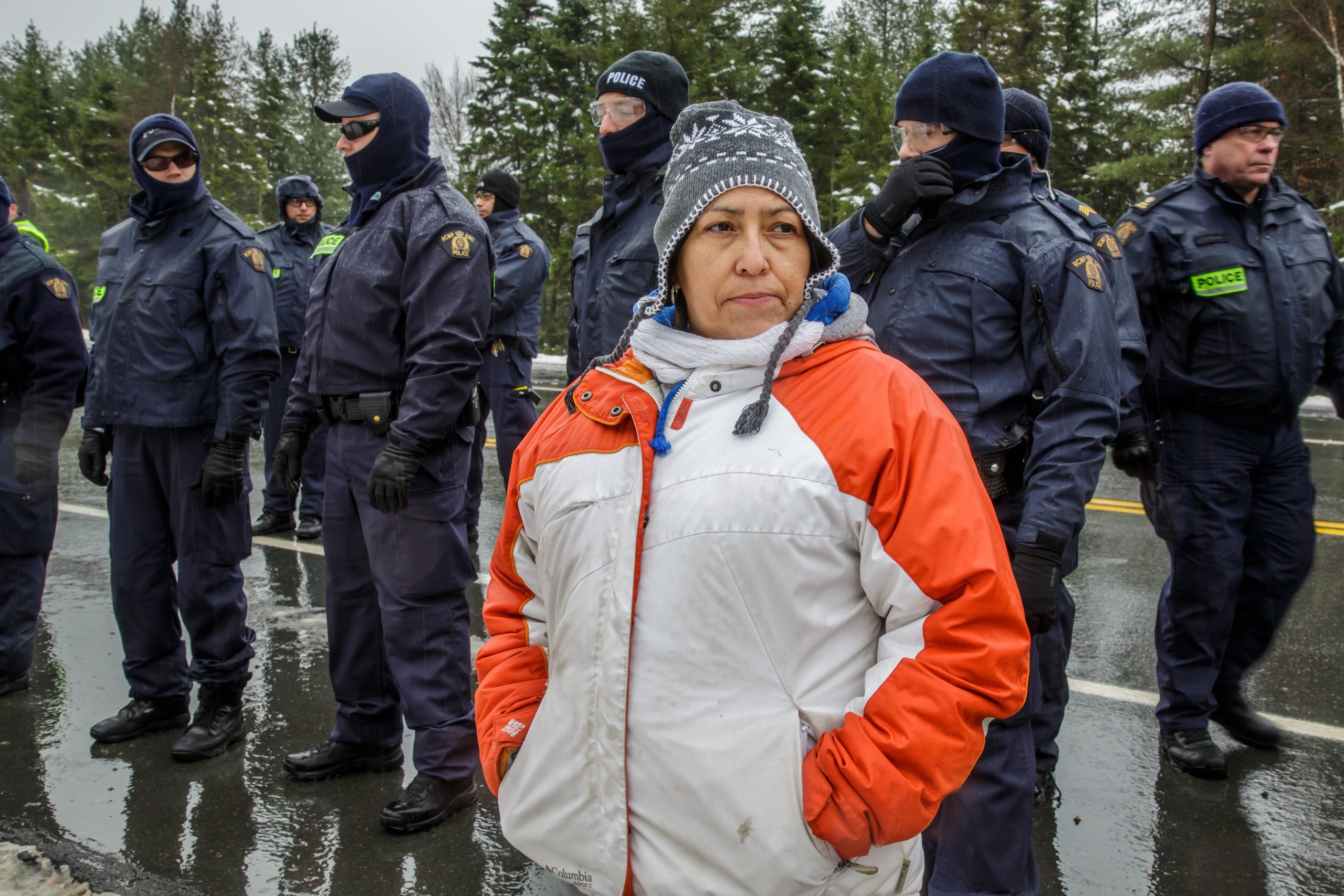 A still from Water Warriors. It shows an Indigenous woman in a winter jacket standing in front of a line of police officers. Her face is contemplative and determined.