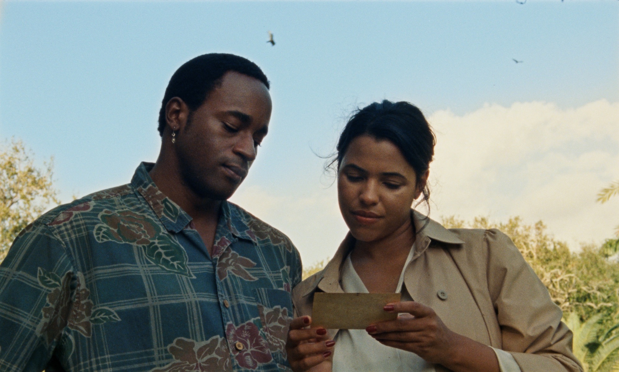 A film still from The Ballad of Suzanne Césaire that shows a Black man and a woman sharing a gaze at a printed photograph she holds. The man, dressed in a floral plaid button-up, and the woman, wearing an open-collared coat with strands of her hair flowing gently in the wind. They are both leaned in close, their eyes locked on the image in thought. They are against a blue sky with a few tree branches peeking out from behind them.