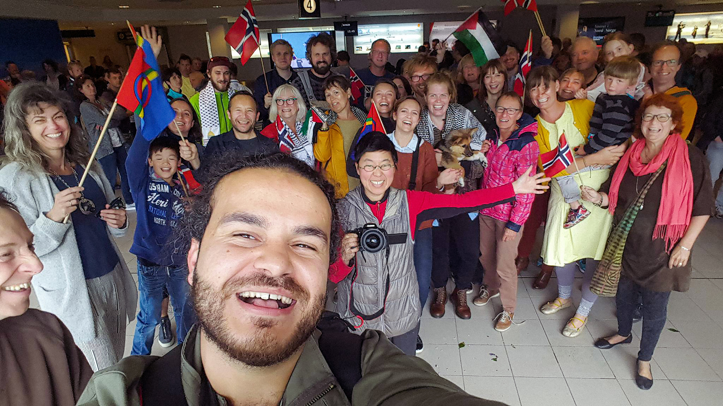 A film still from Life is Beautiful that shows a large group of people in a building gathered in a selfie. They all wear enthusiastic large smiles, some have their shoulders draped with keffiyehs (traditional palestinian scarf) and some hold various flags including the Palestinian flag and Iceland flag.