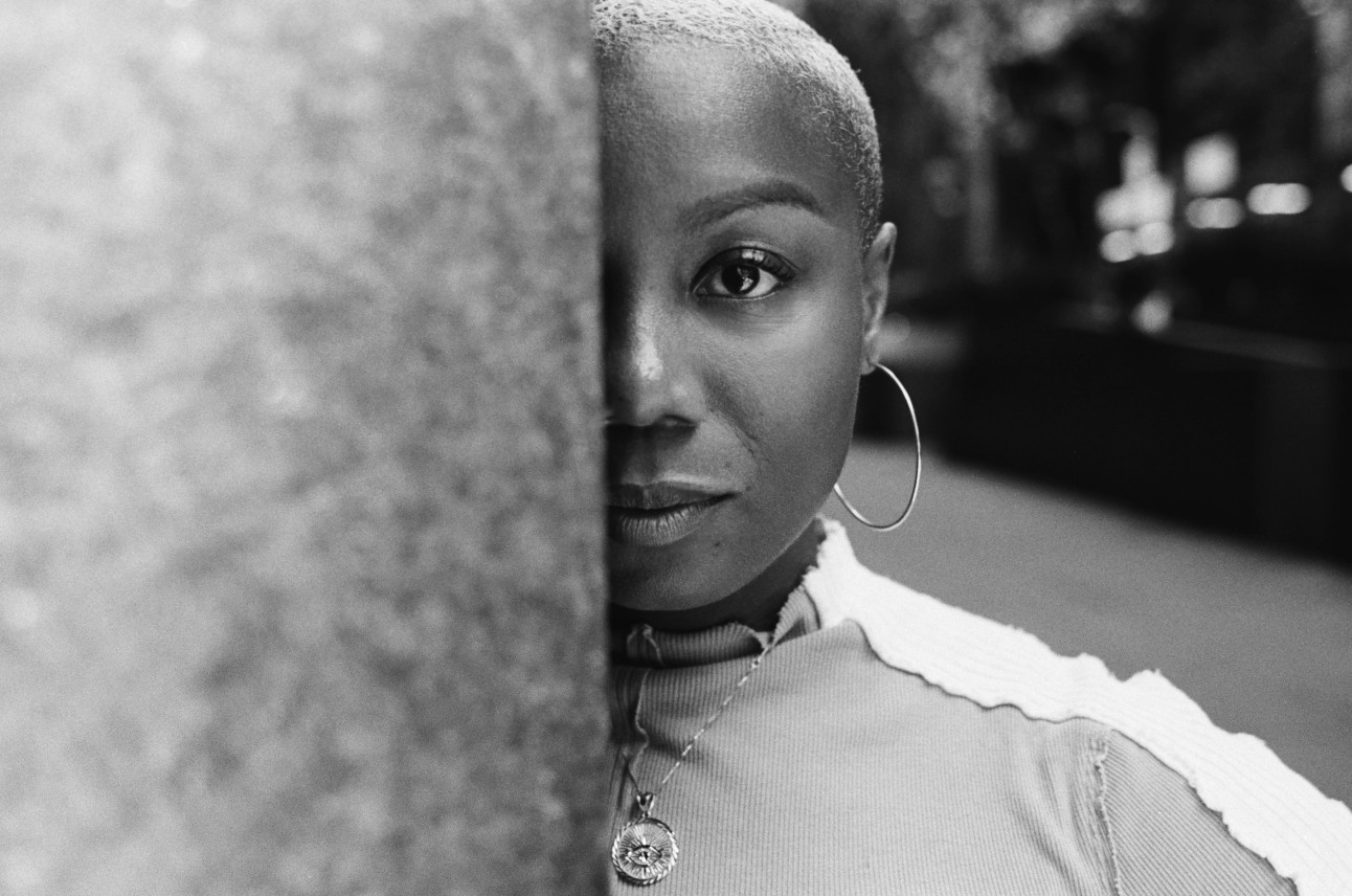 A black-and-white image of Ja'Tovia Gary. She is looking straight ahead at the camera, half of her face is obscured by a wall of some sort. Photo by Gioncarlo Valentine.