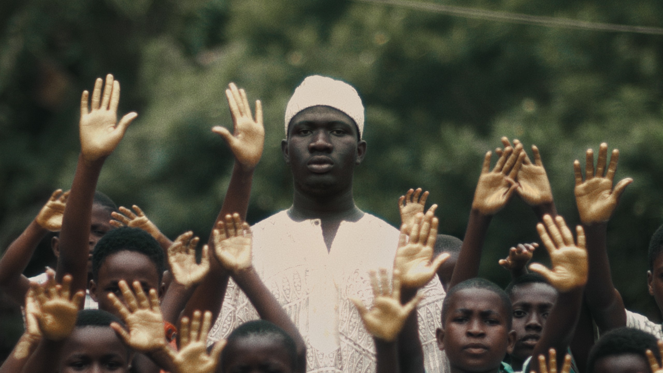 A still from Allumuah shows a Black man dressed in all white standing still, around her are children all with their hands raised in the sky.