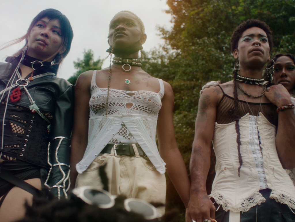 A still from One Magenta Afternoon shows three Black people. They are dressed in mesh clothes and studded necklaces. They are standing outside in a park like setting, the sun illuminates them from behind.