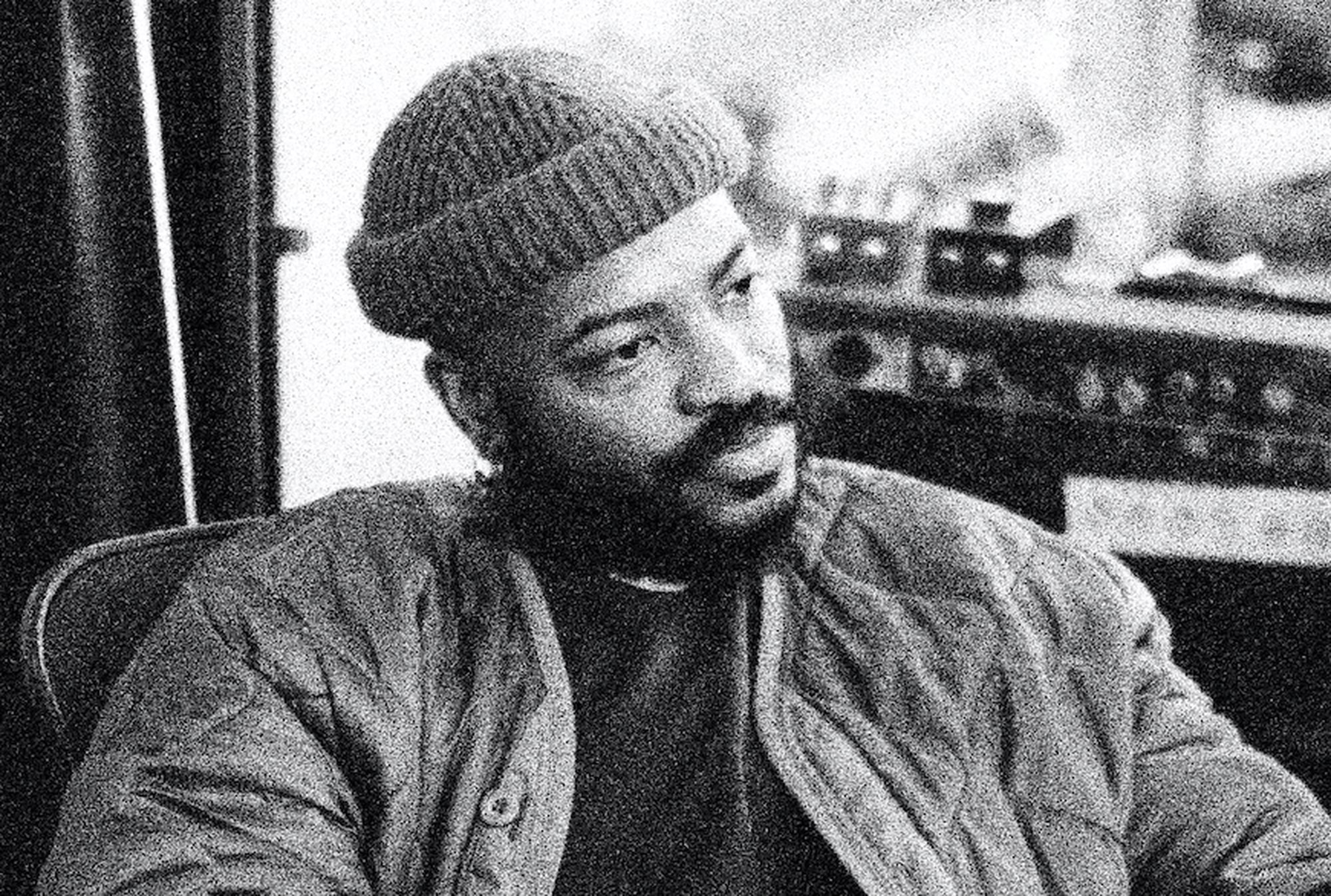 A black-and-white headshot of Darol Olu Kae. He is a black man, he is wearing a beanie and a quilted jacket.