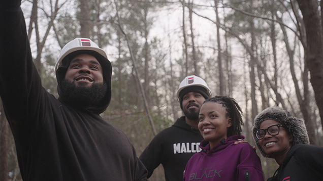 A still from Family Tree shows four Black people standing in a forest. Two are wearing hard hats. They are looking up at something and smiling.
