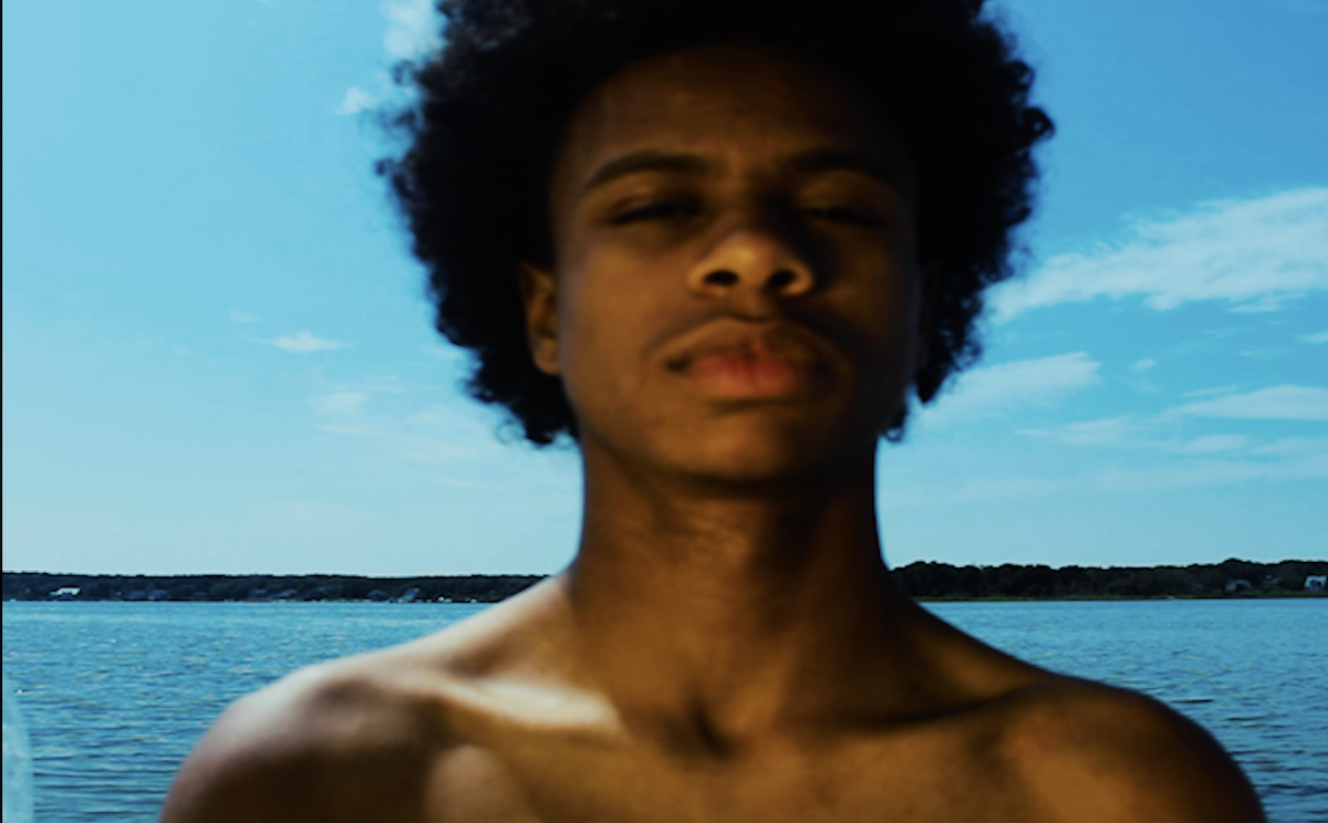 A still shows a man with an afro standing in front of a body of water. His eyes are squinting.