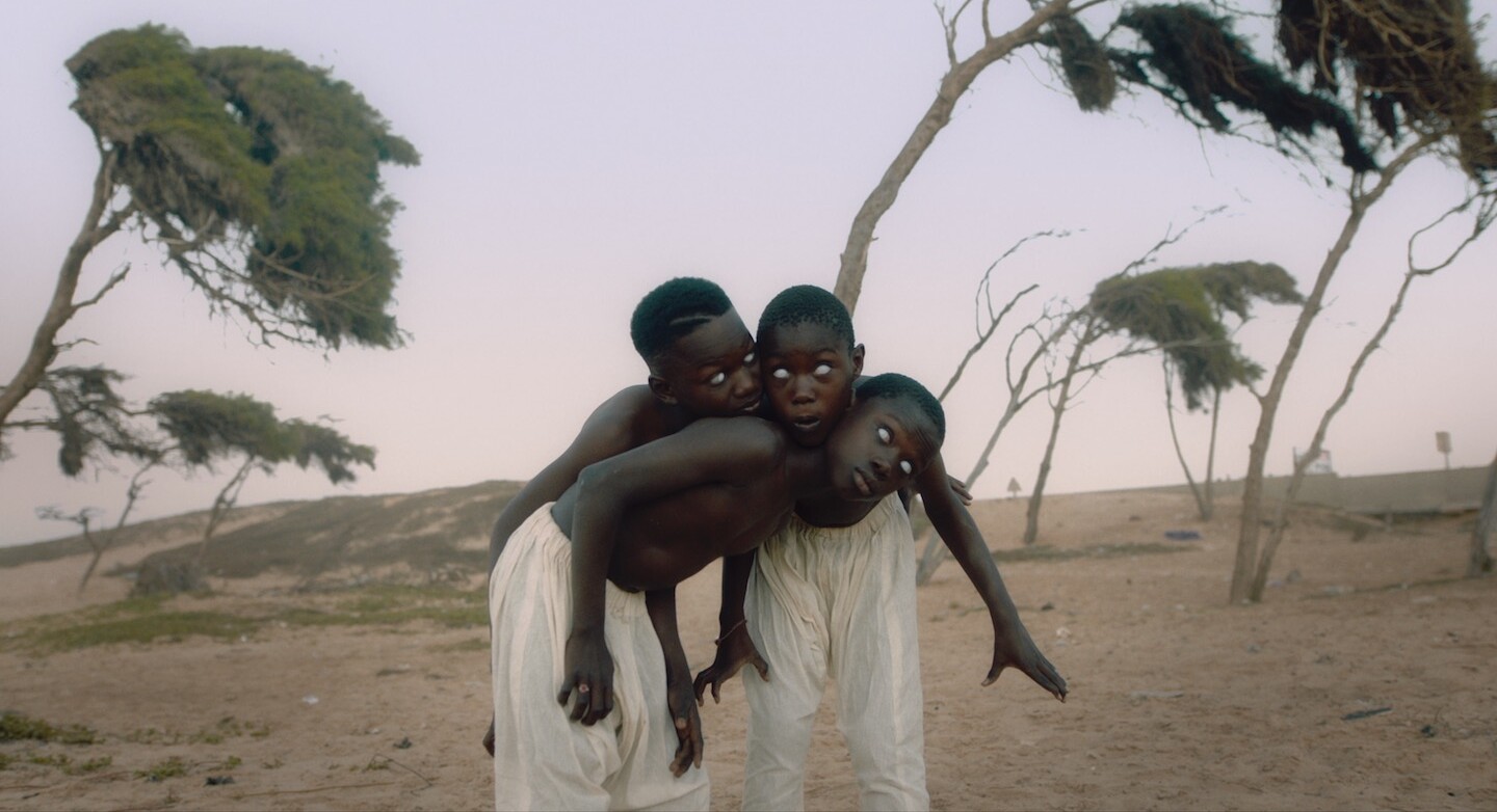 A still from The Dawn shows an arid field. There are three young Black boys standing together, their bodies folded into one another as if they are one. Their eyes are white.