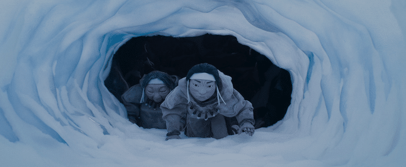 An animated still from The Shaman's Apprentice shows two Indigenous people crawling through a snowy cave. They are wearing large cloaks and have face paint. They have determined expressions.